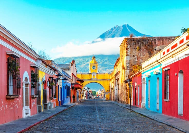 View of Antigua Guatemala's iconic Santa Catalina Arch, with Volcán de Agua towering in the background, framed by colorful colonial buildings lining the cobblestone street.